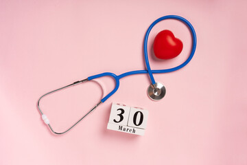 The concept of celebrating Doctor's Day with a stethoscope and a heart on a pink background. Wooden calendar with the date March 30