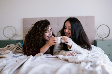two white women lying in bed toasting with a glass of coffee