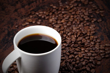 black coffee in white mug with surrounded by roasted coffee beans on dark red wood table