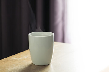 hot coffee in white cup with white smoke blow over on the wood table in a shade of window curtain