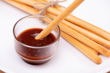 a breadstick dipped with ketchup dipping sauce in transparent cup with pack of breadsticks in background in white dish