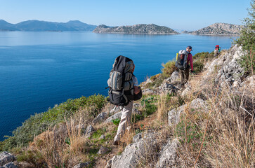 A group of tourists with large backpacks walk along a grassy path along the Mediterranean Sea....