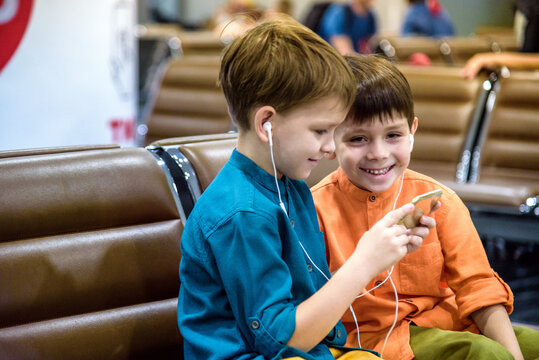 Two Little Boy Sitting In An Airport Departure Hall Contentedly Playing On His Tablet Or Mobile Phone As They Wait For His Flight With His Luggage. Children Are Laughing And Use Earphones.