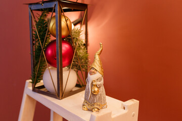 White and golden Santa Claus toy on white shelf on brown background. Red, white and golden baubles and fir tree branch near. Winter holidays, Merry Christmas eve, New Year time concept. Copy space.