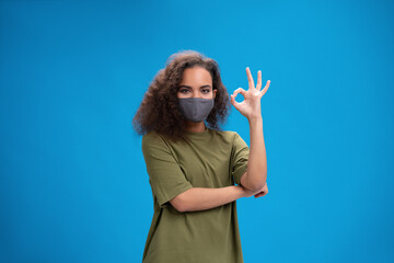 Young African American girl in khaki t-shirt in reusable face mask showing OK gesture, to prevent others from corona COVID-19 and SARS cov 2 infection isolated on pink background. 