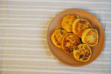 six yellow sugar-free cheesecakes with banana and raisins in corn flour lie in a brown polka-dot round plate on a light white and gray striped tablecloth top view . cooking cheesecakes at home