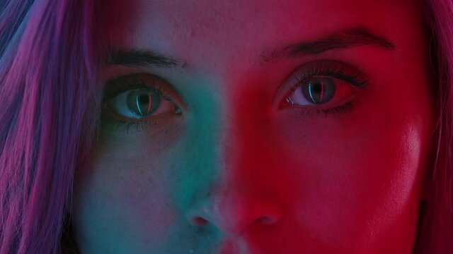Womens eyes with neat natural makeup, illuminated with colorful neon light, look straight ahead. Close up macro portrait. Slow motion.