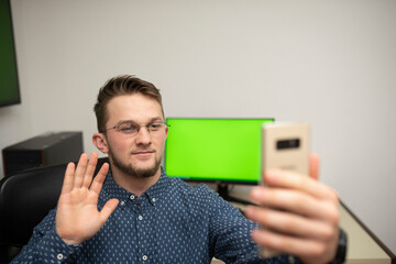 Young caucasian businessman with eyeglasses holding a mobile phone and talking to somebody. Having online meeting. Happy businessman having a good day at job. Computer with green screen in background.