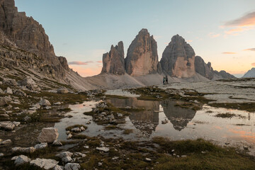 Fototapeta na wymiar A couple enjoying the sunset over the Tre Cime di Lavaredo (Drei Zinnen) mountains in Italian Dolomites. The peaks reflect in a paddle. The mountains are surrounded with orange and pink clouds. Love