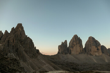 A panoramic capture of the sunset above Tre Cime di Lavaredo (Drei Zinnen) and surrounding mountains in Italian Dolomites. The mountains are surrounded by pink and orange clouds. Golden hour. Serenity