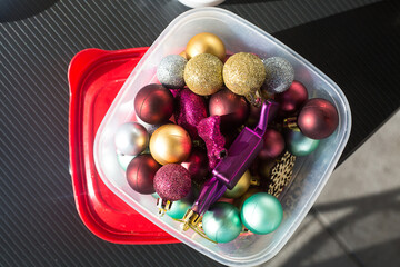 Golden, tiffany and burgundy Christmas balls packed for keeping in a transparent plastic food container on black table. Red cover open. Top view. Storage of new year decoration. Space management