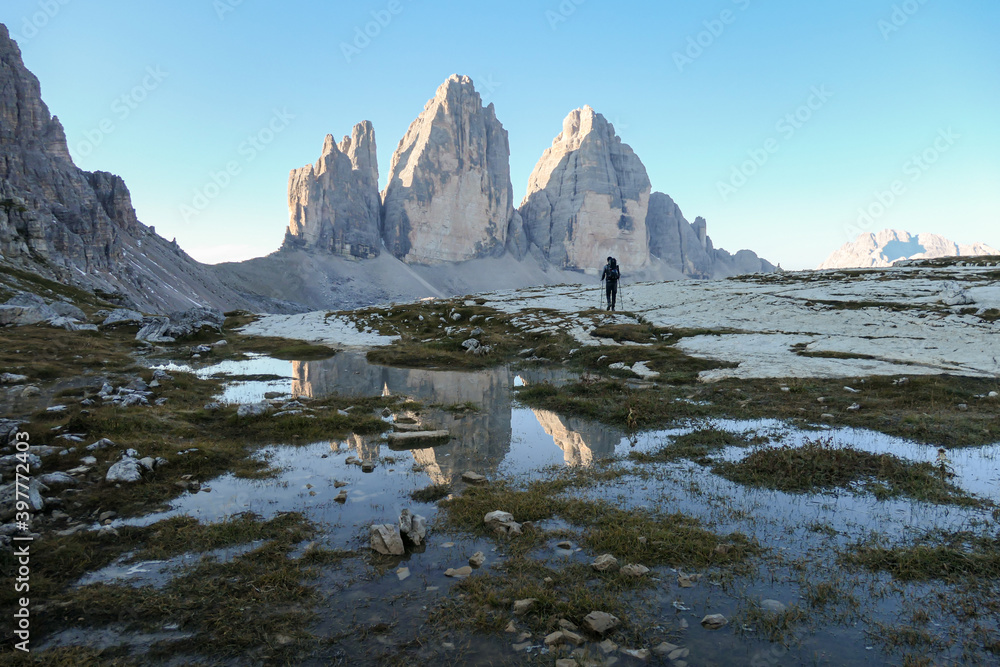 Wall mural A man hiking with the view on the Tre Cime di Lavaredo (Drei Zinnen), mountains in Italian Dolomites. The mountains are reflecting in small paddle. Desolated and raw landscape. Early morning. Daybreak