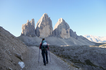 Fototapeta premium A woman in pink jacket hiking on a gravel road and enjoying the view on the Tre Cime di Lavaredo (Drei Zinnen), mountains in Italian Dolomites. Desolated and raw landscape. Early morning. Daybreak