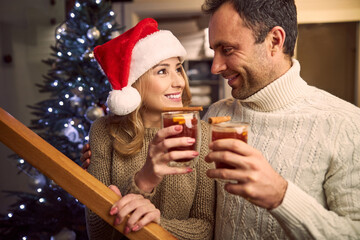 Charming female in Santa hat looking to handsome male in sweater while they holding beverage in arms