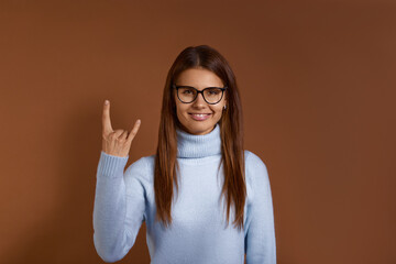 Excited punk young caucasian woman makes rock gesture, looks cool, expresses excitement and happiness, has appealing appearance, wears glasses and light blue sweater, isolated on brown background.