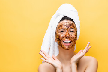 happy beautiful woman with towel on head and coffee mask on face isolated on yellow