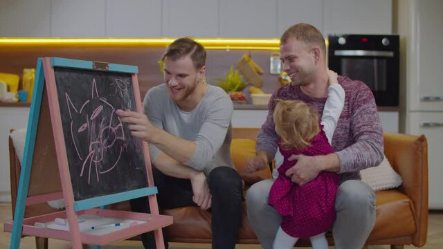 Positive cheerful handsome male lgbt parents with excited cute adopted baby girl drawing picture with colorful chalk on blackboard together, helping kid to develop creative skills in domestic room.