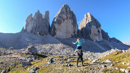 Obraz premium Man carrying woman piggyback with the close up view on the Tre Cime di Lavaredo (Drei Zinnen) in Italian Dolomites. Sharp and high mountain wall. Desolated and raw landscape. Fun and careless moments