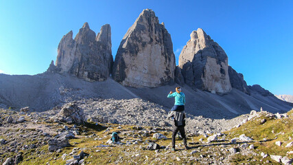 Fototapeta premium Man carrying woman piggyback with the close up view on the Tre Cime di Lavaredo (Drei Zinnen) in Italian Dolomites. Sharp and high mountain wall. Desolated and raw landscape. Fun and careless moments