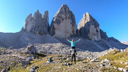 Man carrying woman piggyback with the close up view on the Tre Cime di Lavaredo (Drei Zinnen) in Italian Dolomites. Sharp and high mountain wall. Desolated and raw landscape. Fun and careless moments