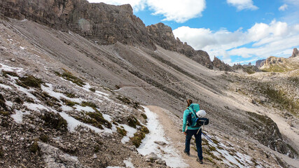 A woman with backpack and sticks hiking on a narrow path in Italian Dolomites. There are sharp and steep mountains around her. Lots of lose stones. Raw and desolated landscape. Following the pathway