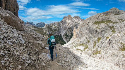 Fototapeta premium A woman with backpack and sticks hiking on a narrow path in Italian Dolomites. There are sharp and steep mountains around her. Lots of lose stones. Raw and desolated landscape. Following the pathway