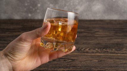 Hand holding glass of brown rum with ice cubes on rustic wooden table with copy-space