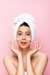 surprised beautiful woman with towel on hair and cosmetic cream on face isolated on pink