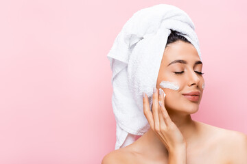 beautiful woman with towel on hair, closed eyes and cosmetic cream on face isolated on pink
