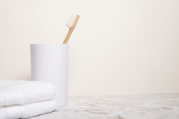 Natural wooden bamboo toothbrush in a white cup next to a white cloth on a marble surface against a magnolia wall with copy space and room for text with left side composition