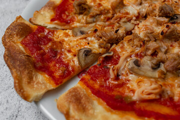 Delicious homemade pizza with tomato, cheese, mushrooms, tuna, vegetables, ham and fresh herbs presented on white plate with light background and selective focus. Copy space.