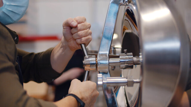Young male worker or owner in apron and mask turning valve on large boiler for beer fermentation