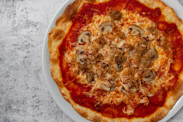 Delicious homemade pizza with tomato, cheese, mushrooms, tuna, vegetables, ham and fresh herbs presented on white plate with light background and selective focus. Copy space.