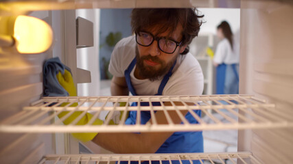 View from inside empty refrigerator of man wearing rubber gloves and apron cleaning shelves