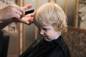 Fototapeta premium A little boy is trimmed in the barbershop. Bright emotions on the face, facial expression, the process of cutting hair.