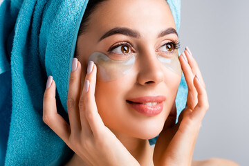 smiling beautiful woman with blue towel on hair and hydrogel eye patches on face isolated on grey