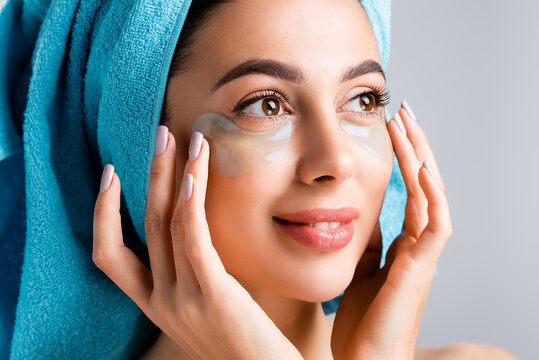 smiling beautiful woman with blue towel on hair and hydrogel eye patches on face isolated on grey