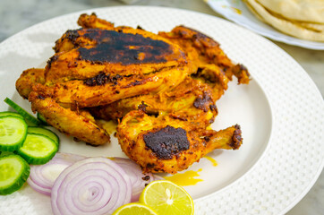 Beautifully placed ready-to-eat full chicken tandoori on a white plate along with sliced onions, cucumbers and lemon