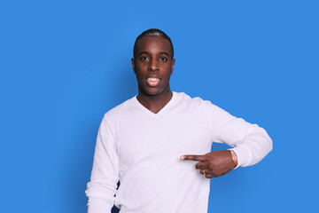 Indoor photo of young African American man pictured isolated on blue background pointing to his white sweater drawing attention to advertisement on it, promoting goods, apps or services