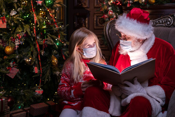 Fototapeta na wymiar Kids and Santa in medical masks. Girl and Santa Claus reading Christmas book sitting near Christmas tree at home. Happy New Year Covid 2021.