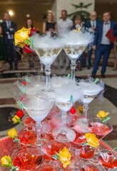 Pyramid of champagne glasses decorated with yellow roses and red cherry fruits, smoke from dry ice