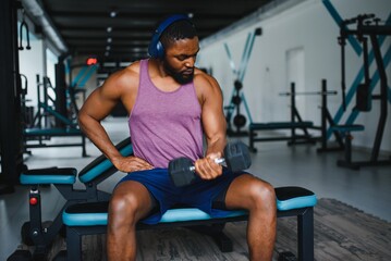 Fototapeta na wymiar Young African American man sitting and lifting a dumbbell close to the rack at gym. Male weight training person doing a biceps curl in fitness center
