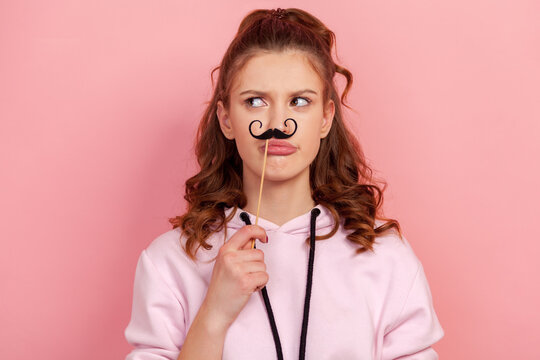 Portrait of funny young female in hoodie holding black paper party mustache on stick, pouting lips and looking to side, having fun on holiday. Indoor studio shot, isolated on pink background