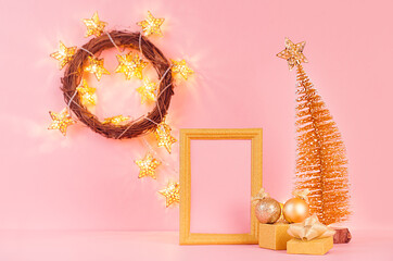 New Year home decor with gold decorations, star lights, christmas tree, wreath, gift box, blank frame for text on soft light pink background.