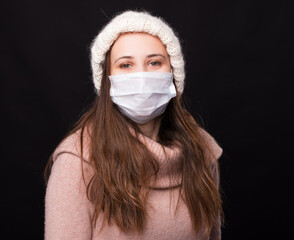 Positive young woman wearing medical face mask, looking right at the camera over black background.
