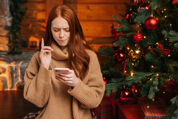 Close-up portrait of pensive redhead young woman wearing winter sweater typing online message using mobile phone on background Christmas tree with bright lighting at living room with festive interior.