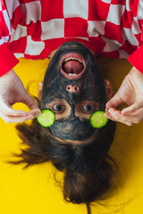 Girl with black mask and cucumbers on her eyes. Relaxing, spa, charcoal mask removes blackheads. Yellow plain background.