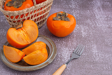 Fresh ripe persimmons with slices of persimmon on a plate with cutlery and whole fruit in a basket with a white brick wall. Space for text. Close-up photo. Concept of health fruit