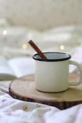 Enamel mug with warm drink and cinnamon stick and open book on the bed. Selective focus.