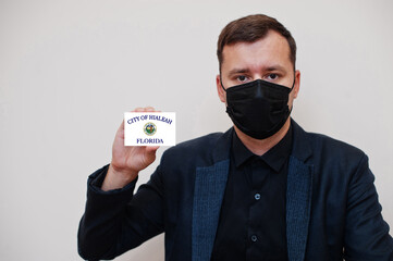 American man wear black formal and protect face mask, hold Hialeah flag card isolated on white background. USA cities coronavirus Covid concept.
